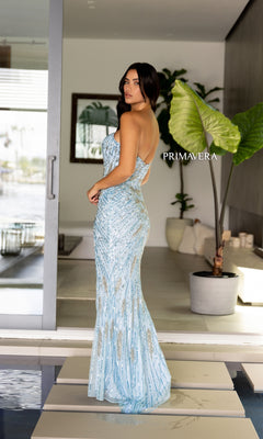 A woman in the Strapless Primavera Long Beaded Formal Dress 4108 stands indoors near a large plant and open sliding glass door. The shimmering light blue evening gown is adorned with sequins.