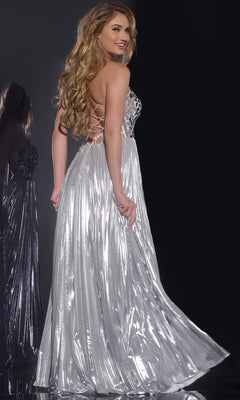 A woman stands against a dark background, looking over her shoulder in the Jovani 41117 Long Prom Dress—a silver metallic, floor-length gown featuring sequin embellishments and a lace-up open back, with long wavy hair cascading down.