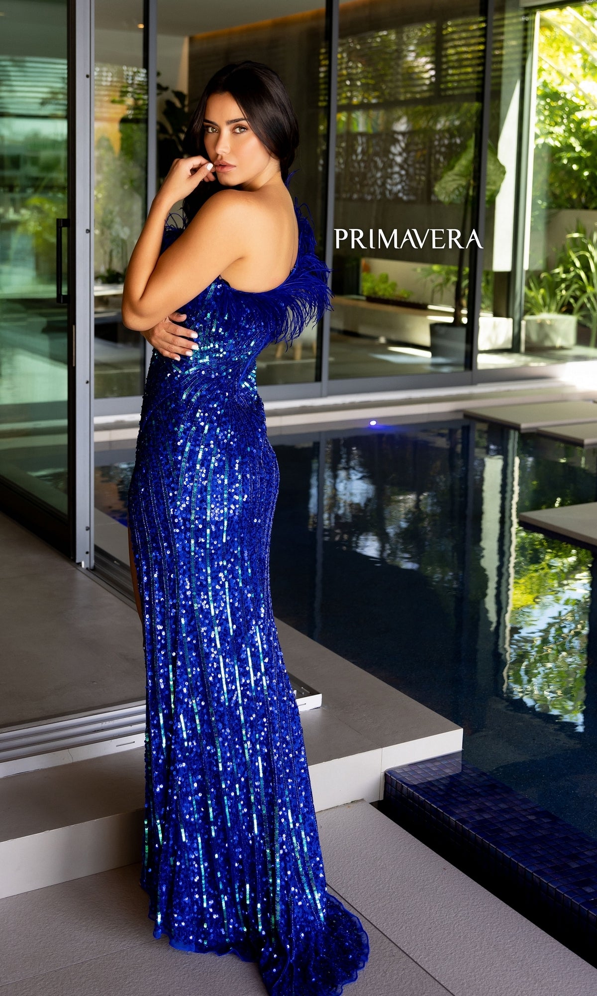 A woman in a one-shoulder, blue Primavera Feathered Long Sequin Prom Dress 4112 stands by an indoor pool, looking over her shoulder. The word 