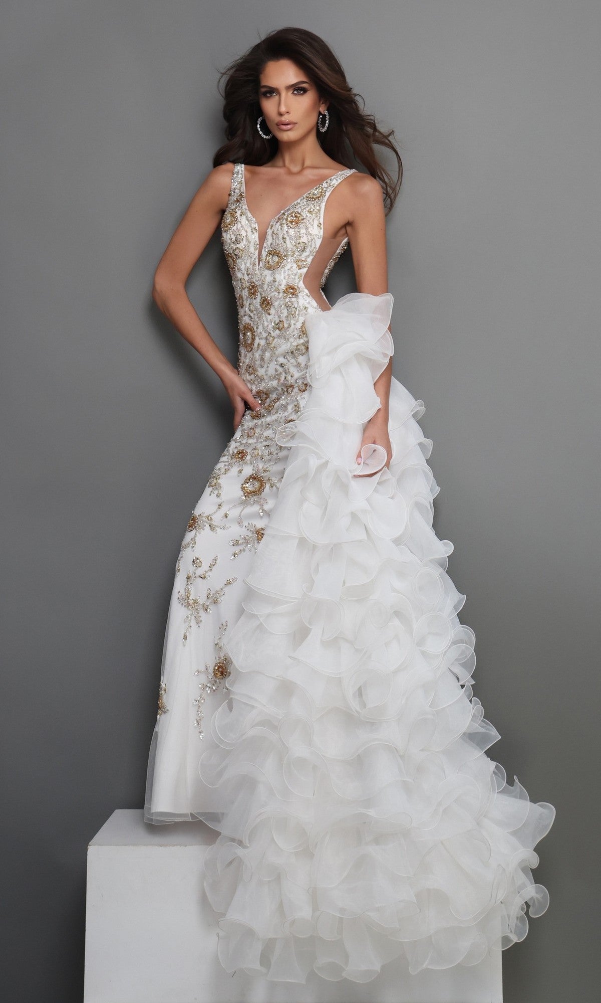 A woman wearing the Long Formal Dress: Jovani 41146 in off-white, a fitted designer gown adorned with elegant gold embroidery and intricate gold beadwork, featuring a dramatic ruffled mermaid silhouette skirt, poses against a plain gray background.