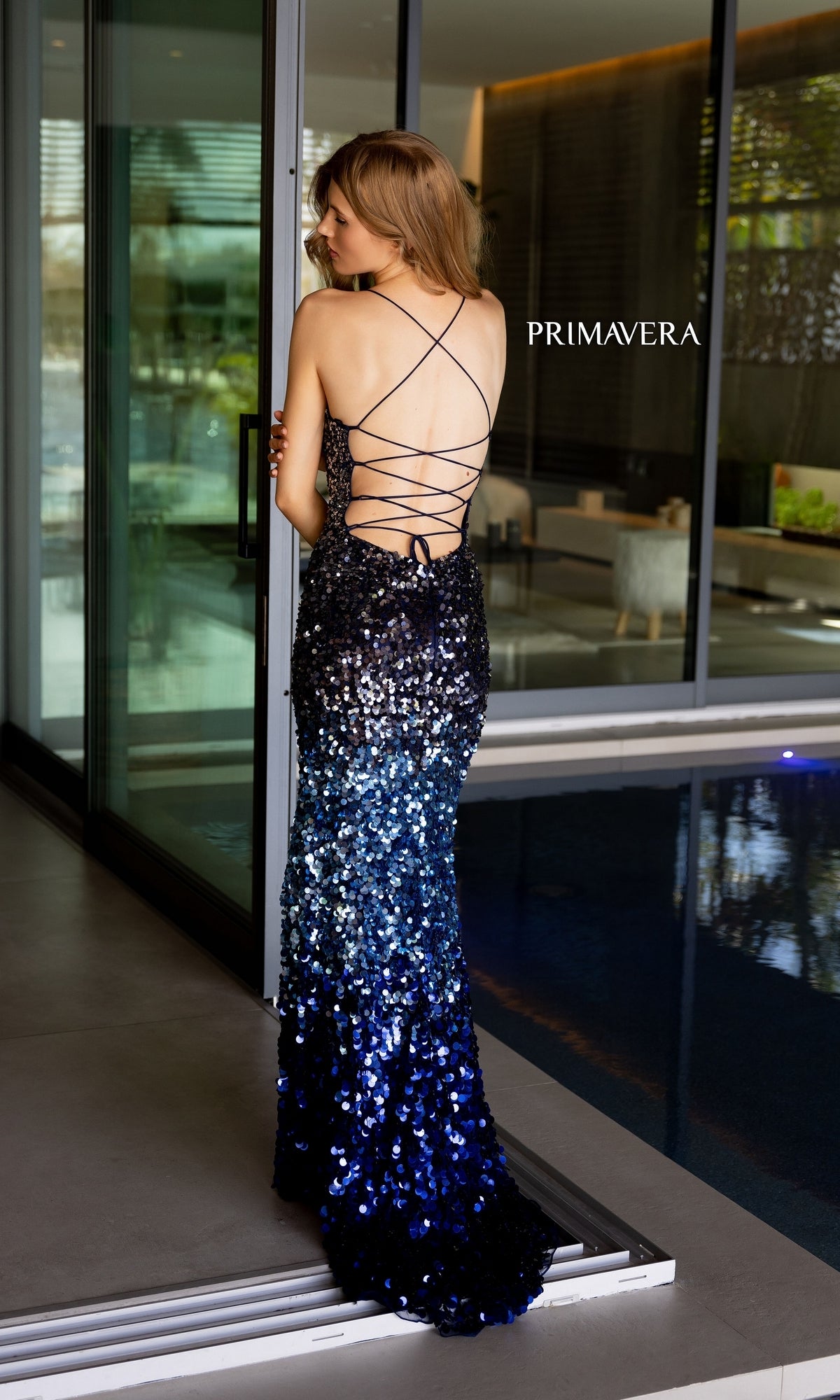 A woman wearing the Primavera Backless Long Ombre Sequin Prom Dress 4114 with crisscross straps stands near a glass door by the pool. 