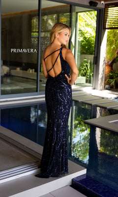 A woman stands by an indoor pool, facing away, wearing the Primavera One-Shoulder Beaded Formal Gown 4118 in navy blue with shimmering sequins and a strappy open back. "PRIMAVERA" is displayed on the wall.