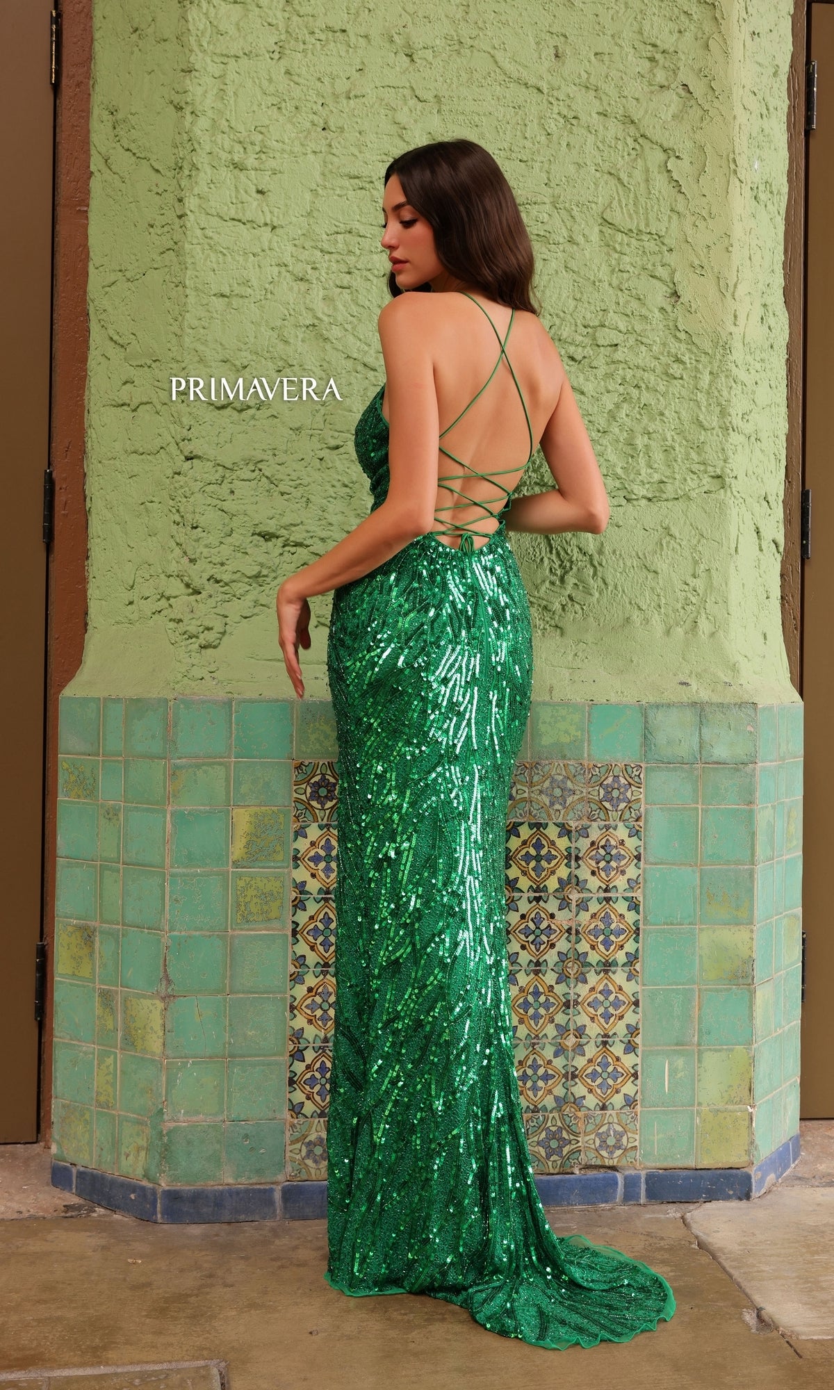 A woman stands with her back to the camera in the Primavera Beaded-Print Long V-Neck Prom Dress 4121, its backless formal design and crisscross straps shimmering against a green wall and tiled floor.