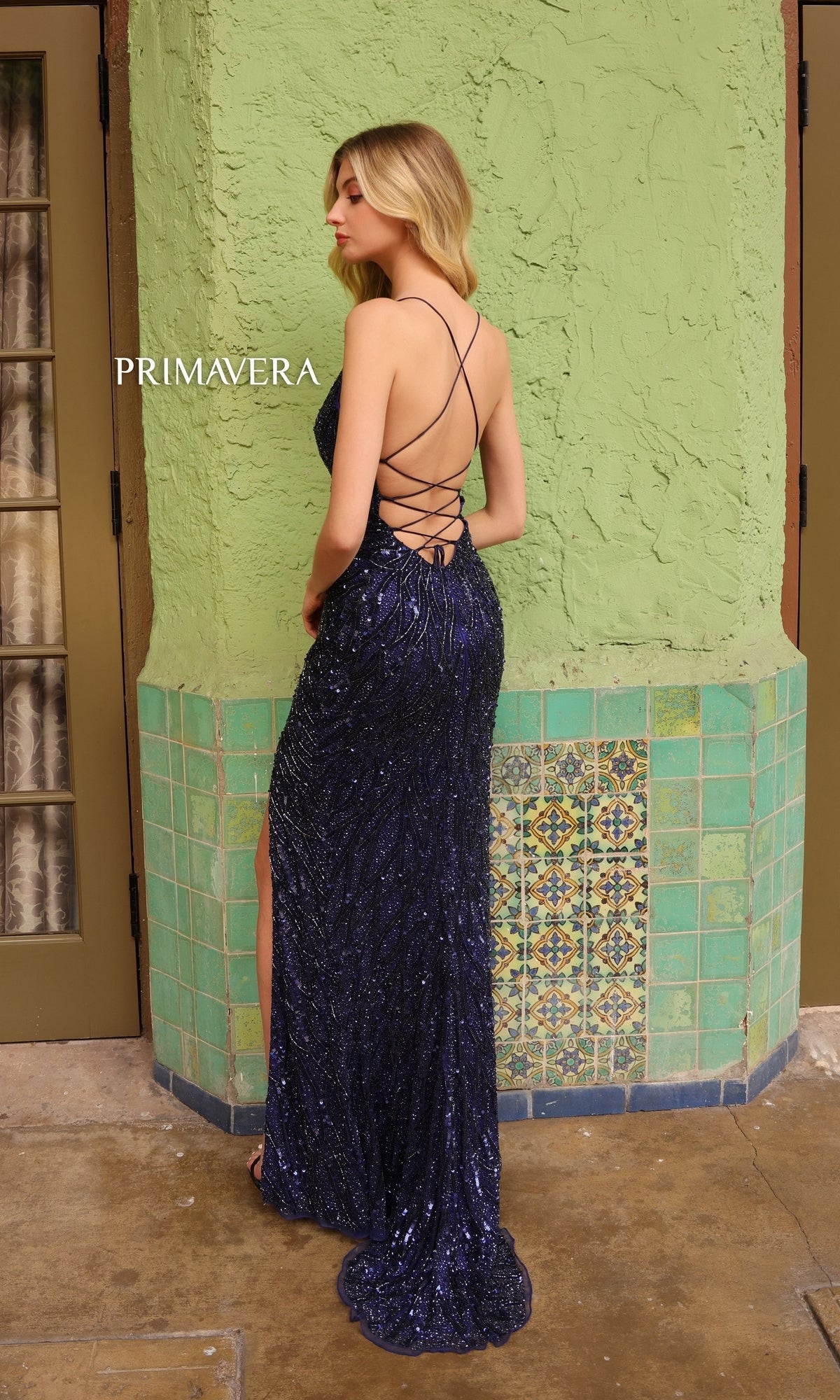 A woman in the Primavera Beaded-Print Long V-Neck Prom Dress 4121 stands by a green tiled wall, showcasing its dark blue sequins, open back, and crisscross straps.