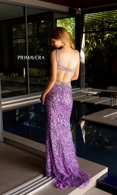 A woman stands by a pool wearing the Primavera One-Shoulder Beaded Long Prom Dress 4126. The floor-length, sleeveless lavender gown features sparkling beadwork, and "PRIMAVERA" is visible on a nearby window.