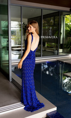 A woman in a sleeveless Beaded-Fringe Long Primavera Prom Dress 4129 with an open back stands by an indoor pool. The floor-length blue gown’s elegance is highlighted, with "PRIMAVERA" illuminated in the background.