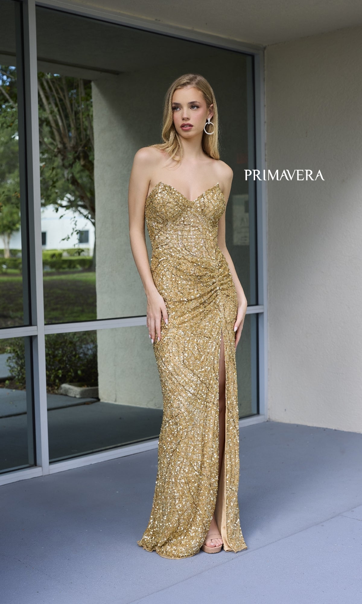 A woman stands outside near a window in the Primavera Sheer-Bodice Beaded Long Prom Dress 4130, a strapless gown with gold sequins and a thigh-high slit. The word 