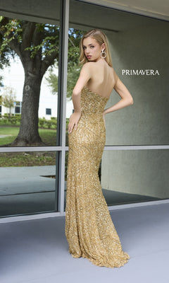 A woman radiates elegance outdoors in the Primavera Sheer-Bodice Beaded Long Prom Dress 4130, a strapless gown featuring gold sequins, posing by large windows with trees and grass in the background.