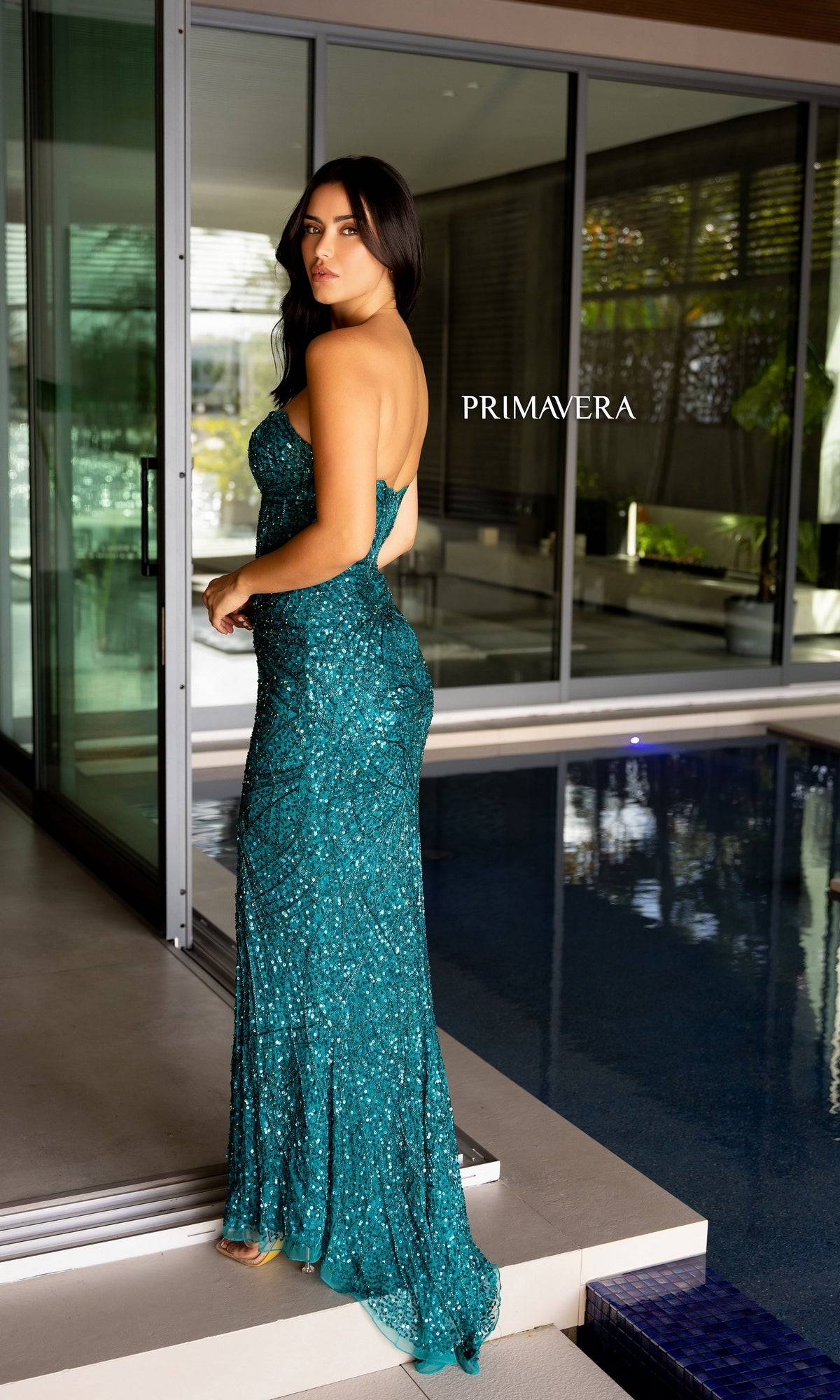 A woman in the Primavera Sheer-Bodice Beaded Long Prom Dress 4130, shimmering with sequins, stands by an indoor pool and gazes over her shoulder. 