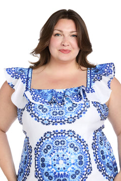A woman with medium-length brown hair wears the R&M Richards 4134W plus-size short party dress, featuring a blue print and ruffled sleeves, while standing against a white background.