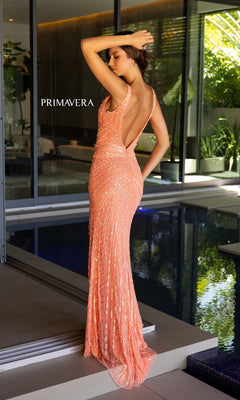 A woman in the Primavera Long Beaded Designer Prom Dress 4143 stands by an indoor pool, facing away with one arm raised, near large windows and lush tropical plants.
