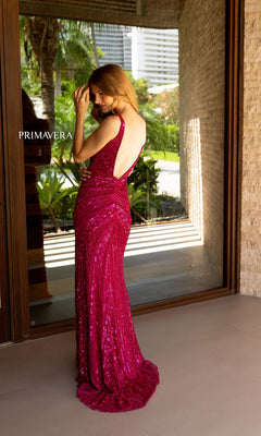 A woman wearing the Primavera Long Beaded Designer Prom Dress 4143 in sparkly deep pink stands indoors by a glass door, with green foliage outside.
