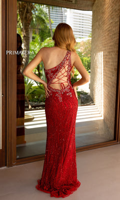 Wearing the Primavera One-Shoulder Beaded Long Prom Dress 4144, a woman stands with her back to a glass door, framed by tropical plants and buildings in the background.