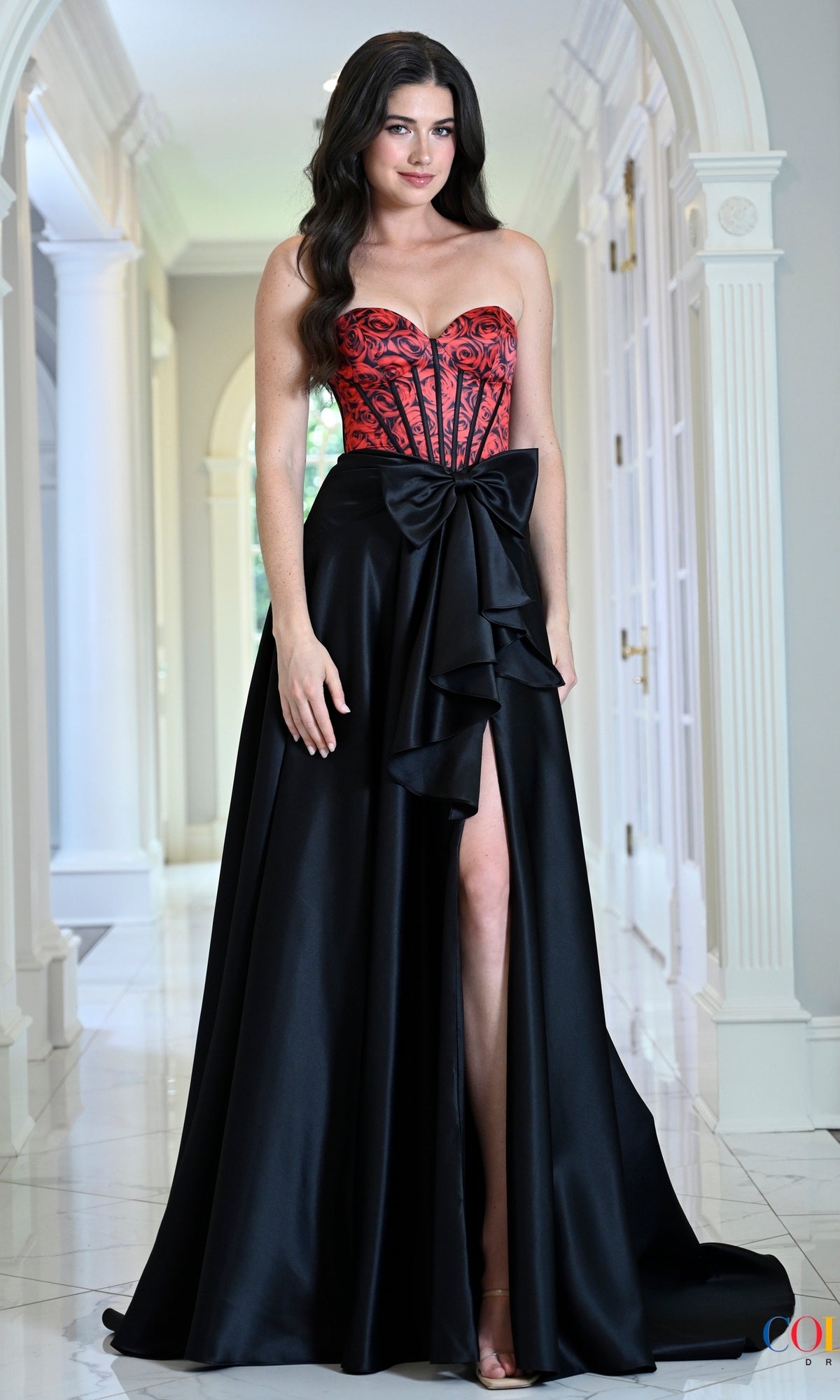 A woman wears a Colors Dress 4149 gothic prom dress featuring a red corset bodice with a black rose pattern and a floor-length skirt with a front slit and bow detail.