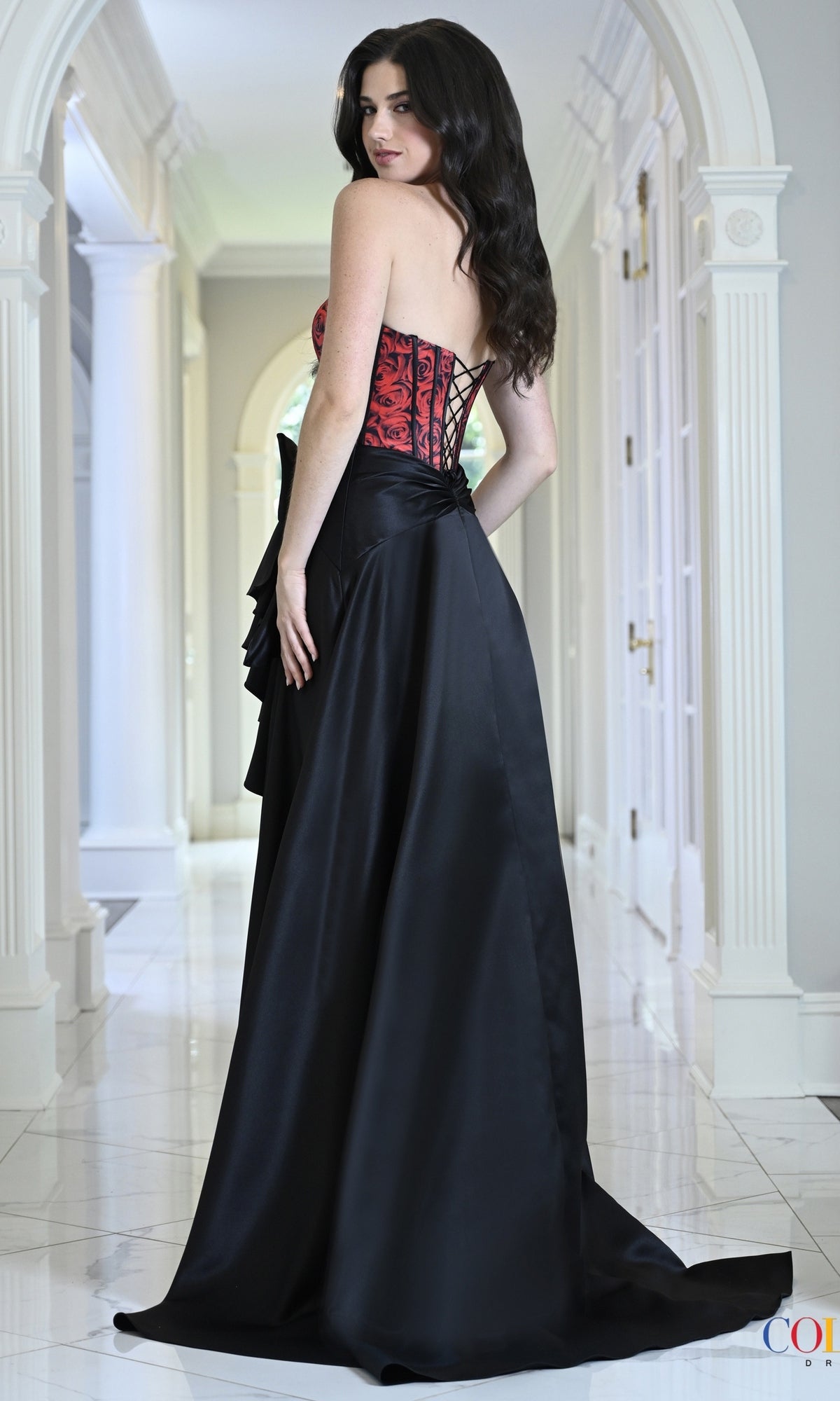A person wearing a Colors Dress 4149 long prom dress, featuring a rose-patterned corset bodice and a black bustle skirt.