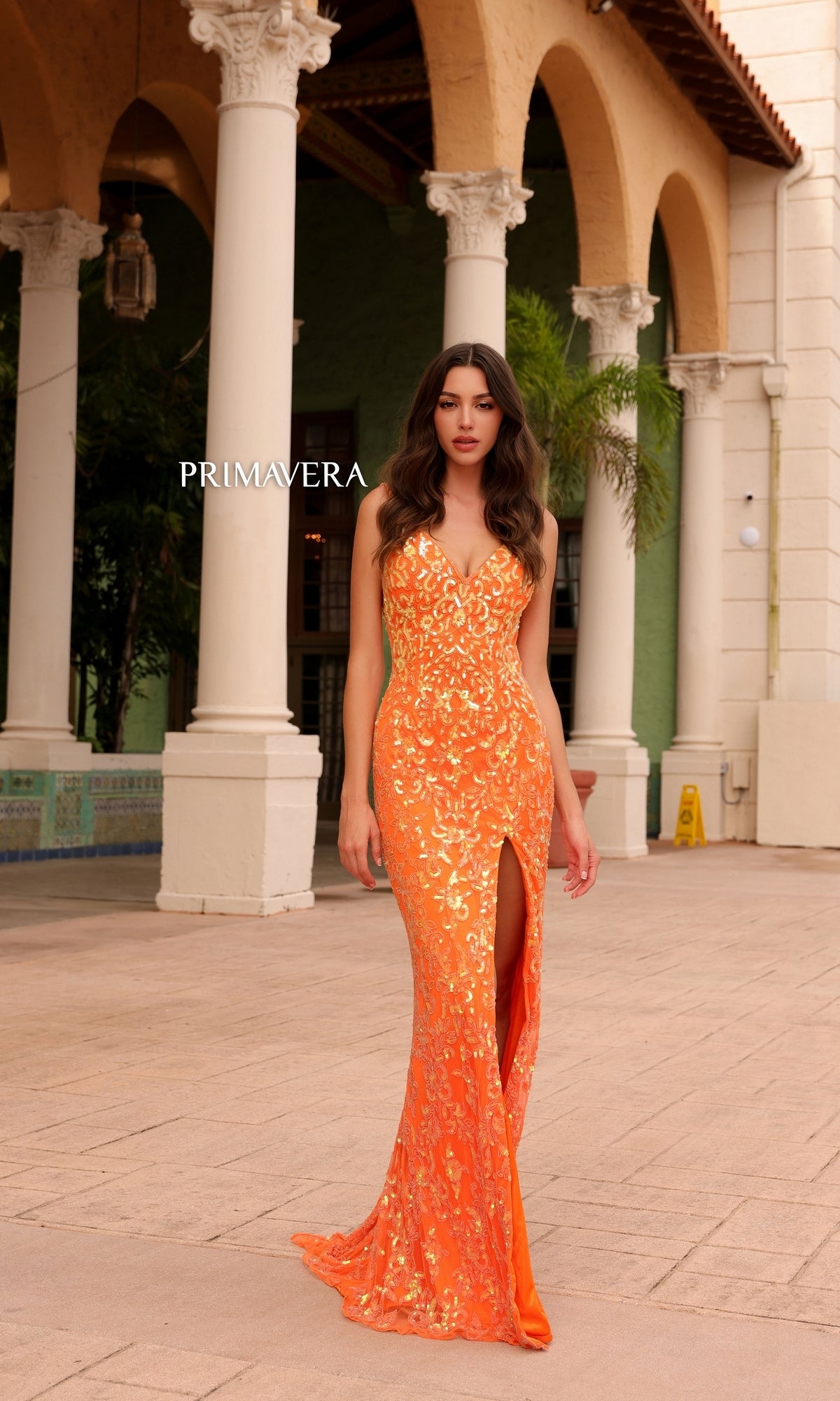 A woman in the Primavera Ombre-Beaded Long Formal Gown 4150, a fitted orange strapless dress with a front slit, stands before columns and arches with 