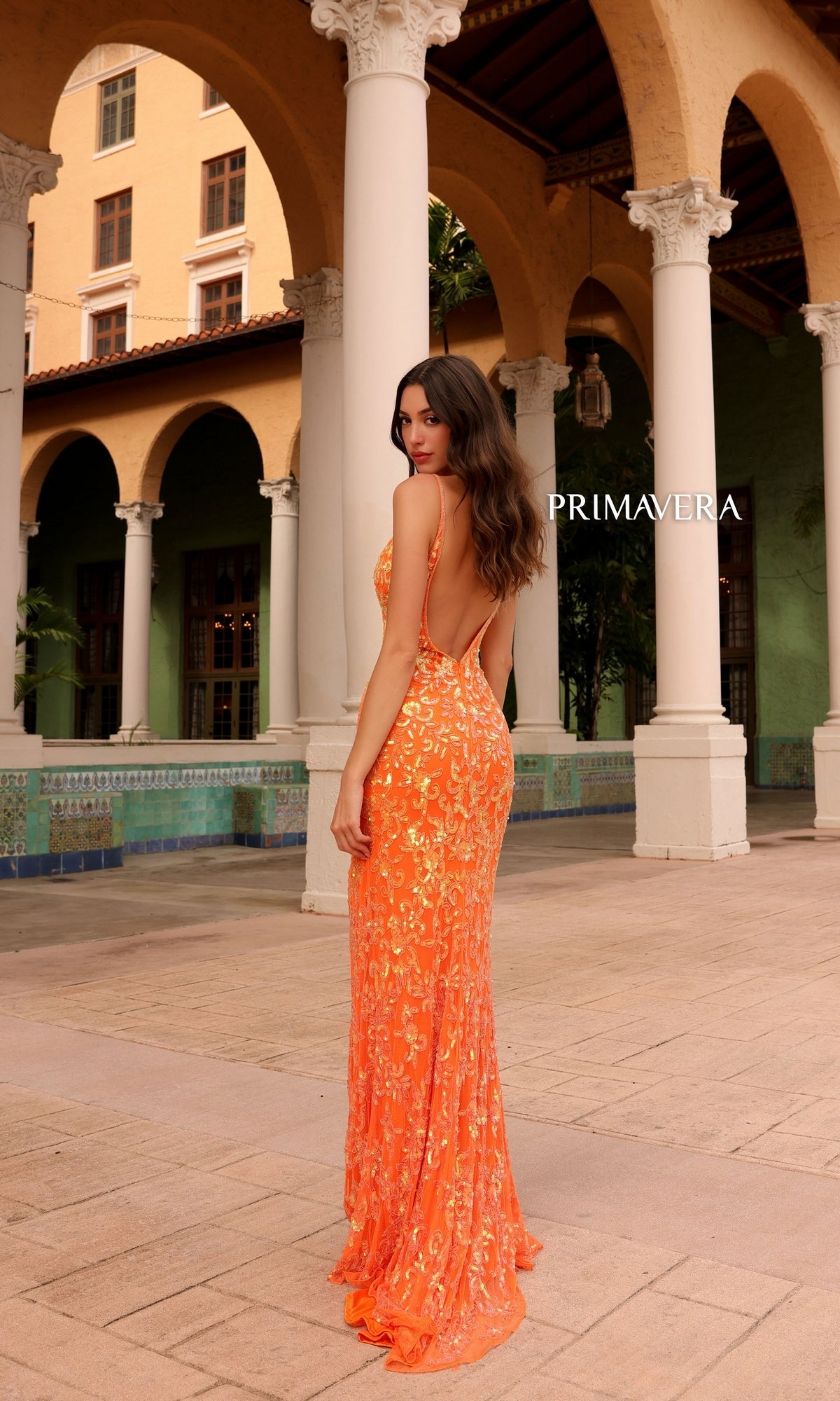 A woman in the Primavera Ombre-Beaded Long Formal Gown 4150, featuring a backless design and orange sequin ombre detailing, stands outside by columns and arches of a Mediterranean-style building with 