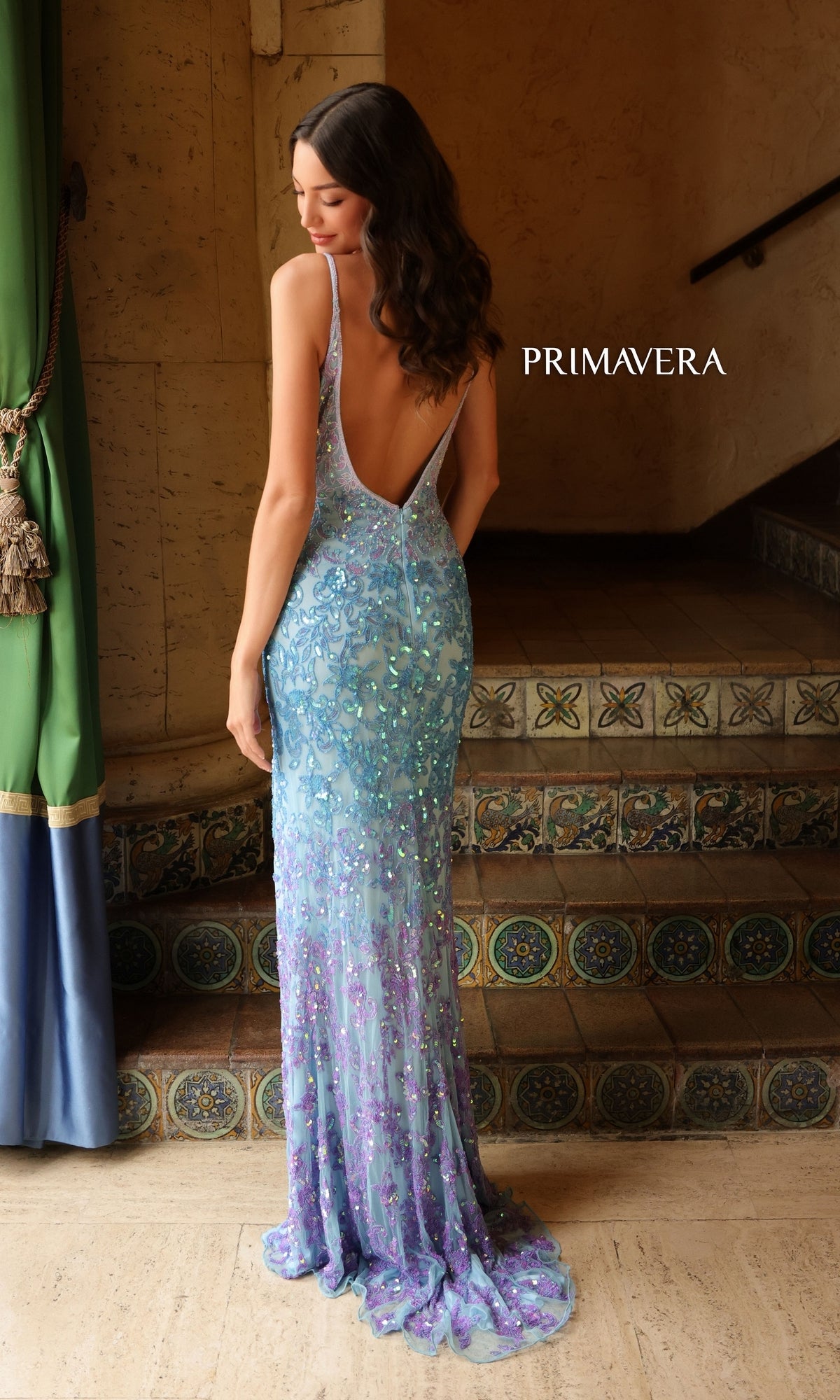 A woman stands on tiled steps, facing away, in the Primavera Ombre-Beaded Long Formal Gown 4150—its sparkling light blue and purple beads glimmer. 