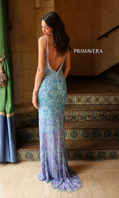 A woman stands on tiled steps, facing away, in the Primavera Ombre-Beaded Long Formal Gown 4150—its sparkling light blue and purple beads glimmer. "PRIMAVERA" appears on the wall beside her.