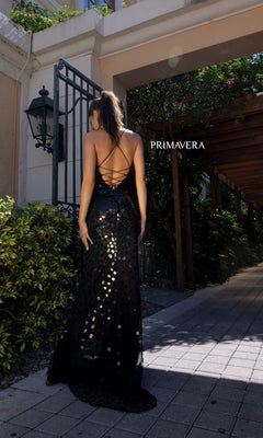 A woman stands outdoors on a stone path, facing away, wearing the Primavera Broken-Glass Long Formal Dress 4151—black, floor-length, backless, and accented with sparkling sequin details. "PRIMAVERA" is visible in the background.