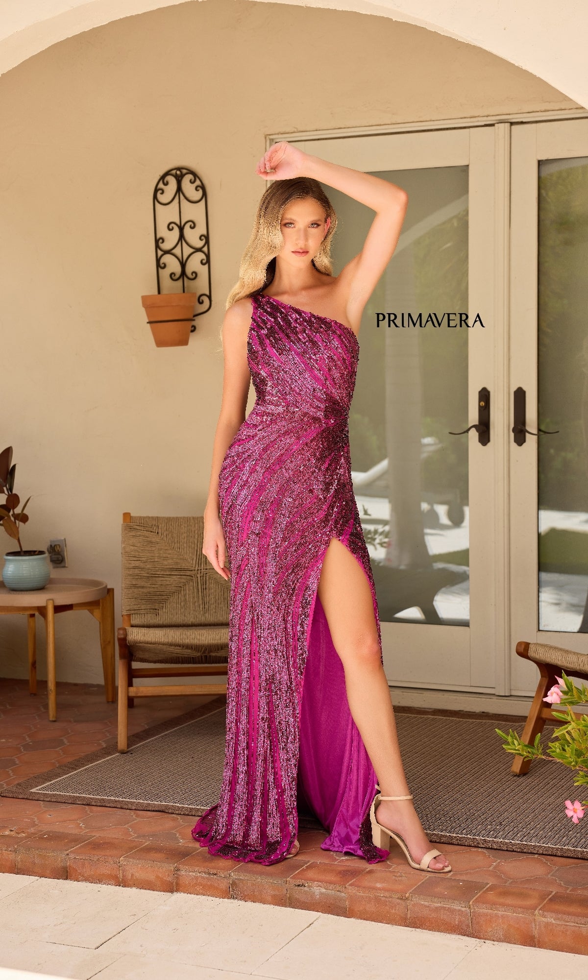 A woman stands on a porch in front of glass doors, wearing the glamorous Primavera One-Shoulder Long Beaded Prom Dress 4152—a magenta, sequined gown with a thigh-high slit. The word 