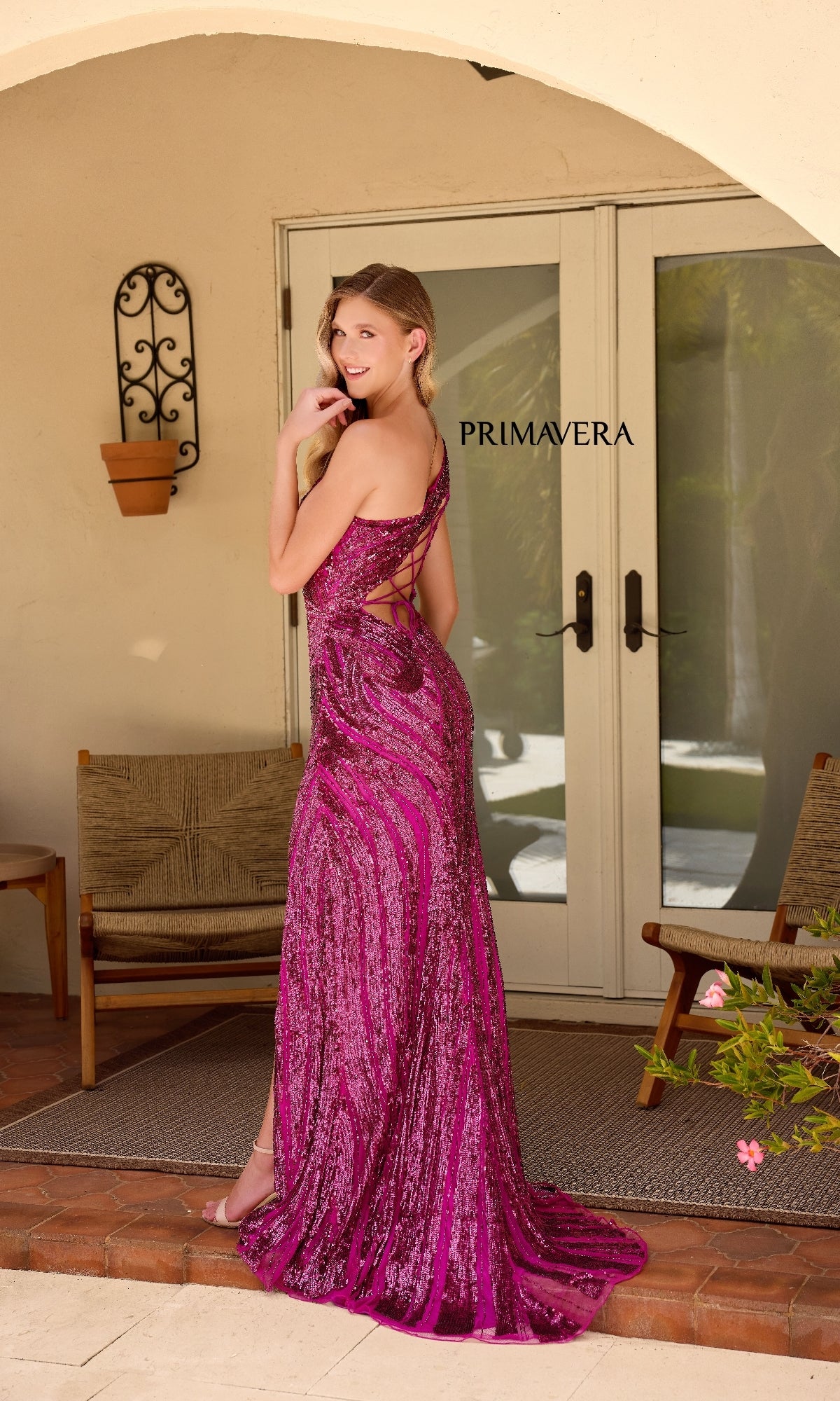 A woman in a sparkling magenta Primavera One-Shoulder Long Beaded Prom Dress 4152 stands on a patio, smiling as she faces away, with 