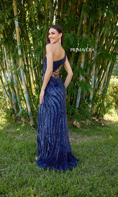 A woman stands outdoors before bamboo, smiling over her shoulder in the Primavera One-Shoulder Long Beaded Prom Dress 4152 featuring a blue sequin design, open back, and elegant lace-up detail.
