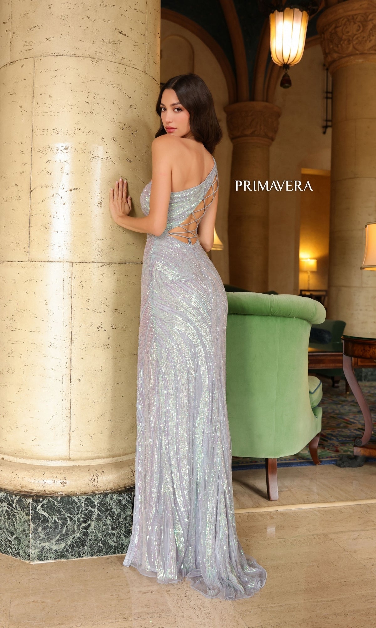 A woman in a shimmering, light purple Primavera One-Shoulder Long Beaded Prom Dress 4152 with a crisscross back stands by a marble column in an elegant, decorated lobby.