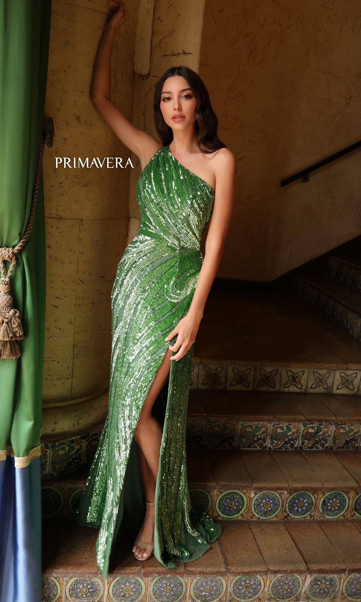 A woman poses on tiled stairs in the Primavera One-Shoulder Long Beaded Prom Dress 4152, a stunning green sequined gown with a thigh-high slit and elegant one-shoulder design, perfect for prom night.