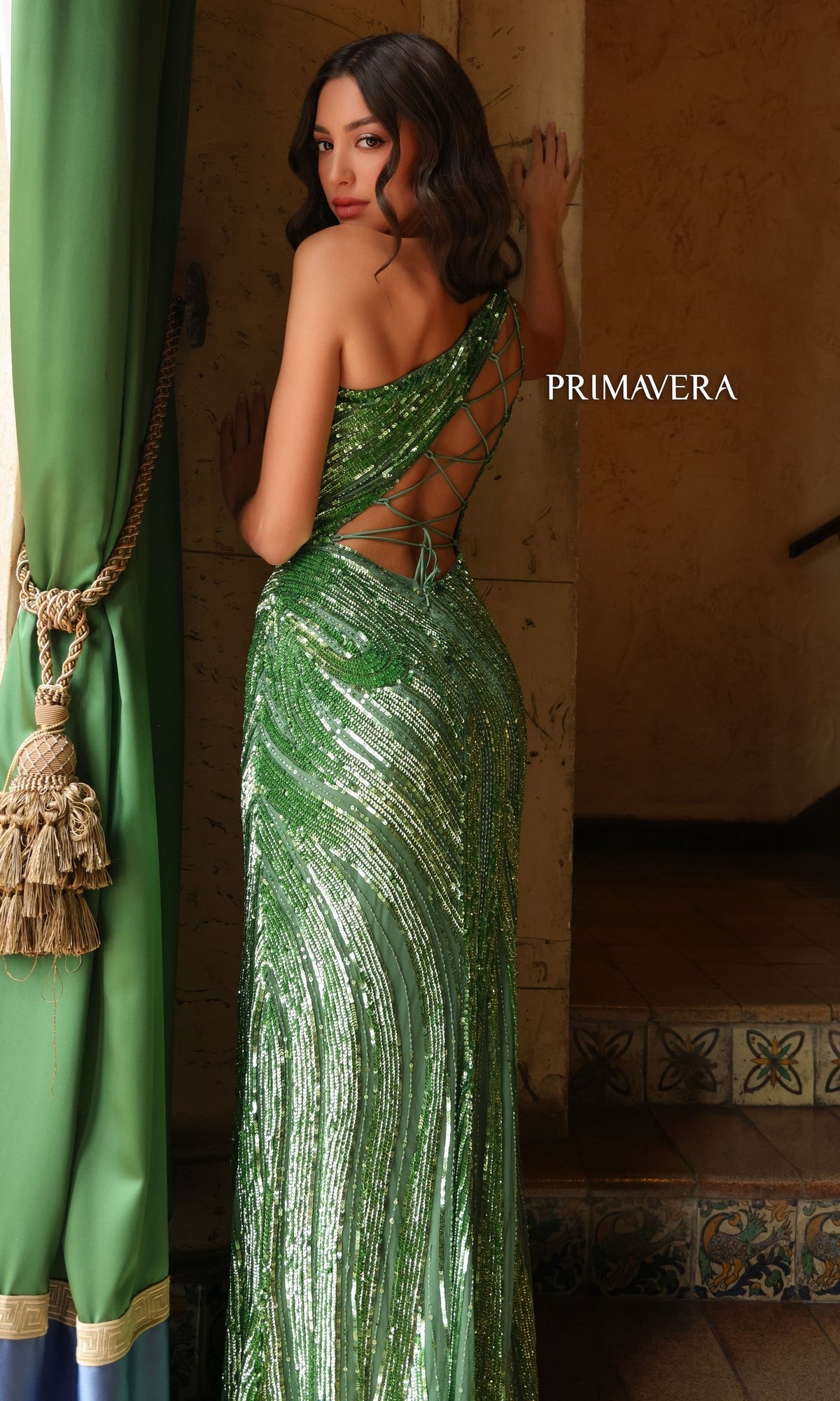 A woman in the Primavera One-Shoulder Long Beaded Prom Dress 4152 stands by a curtain, facing away to show the lace-up back. The word “PRIMAVERA” appears on the wall beside her.