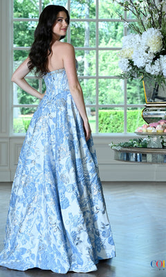 A woman viewed from the back wearing a Colors Dress 4153, a floor-length floral jacquard prom dress with a blue and silver pattern.