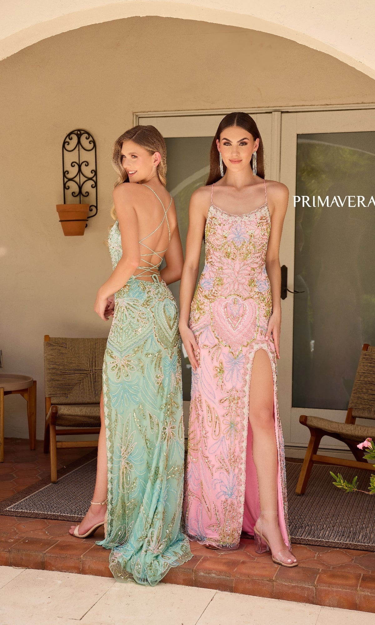 Two women pose side by side on a patio wearing Primavera 4156 beaded-print designer prom dresses—one in light blue with a lace-up back, the other in pink with a thigh-high slit—showcasing elegant style.