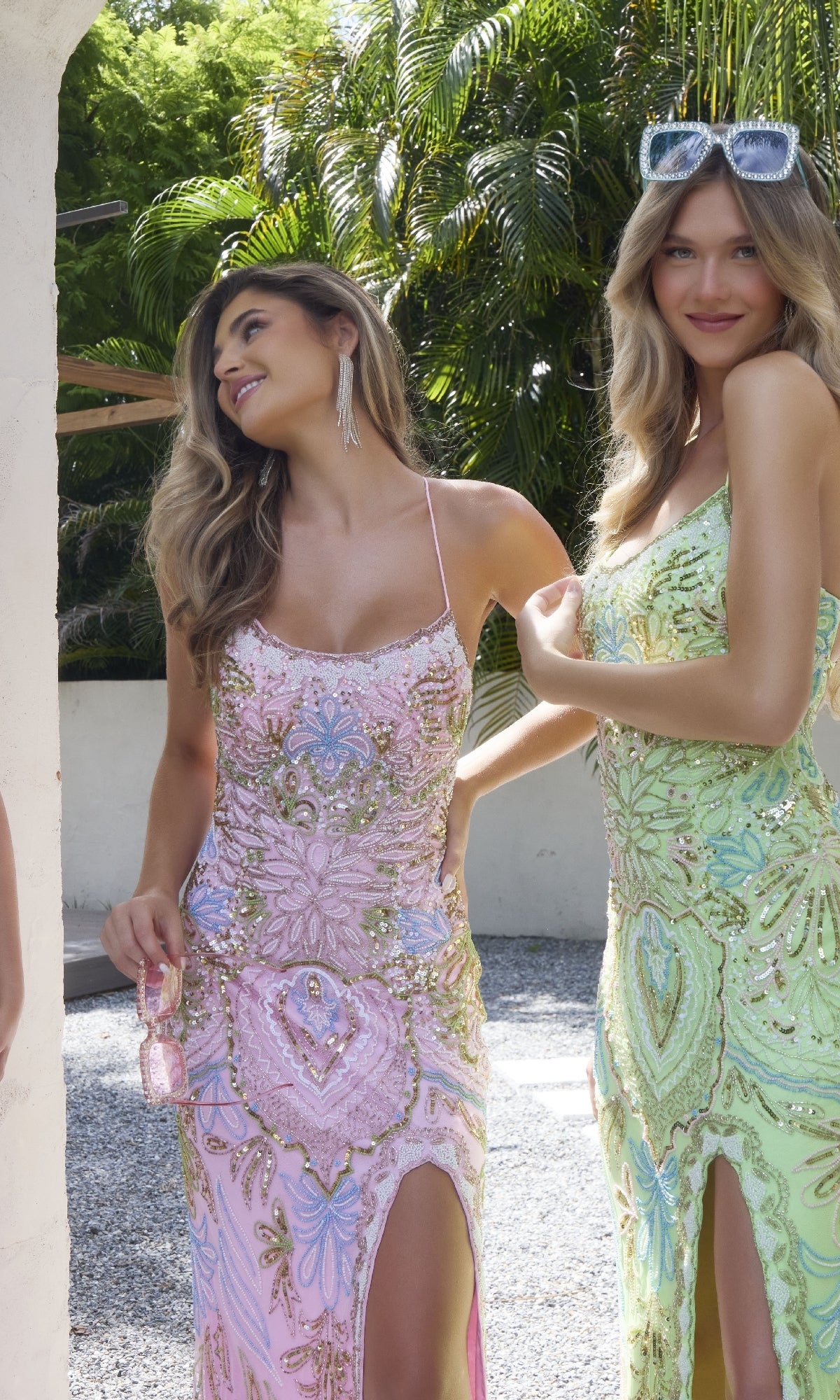 Two women stand outdoors in Primavera 4156 beaded-print designer prom dresses with pastel patterns and thigh-high slits; one wears pink and holds a purse, the other wears green with sunglasses on her head.