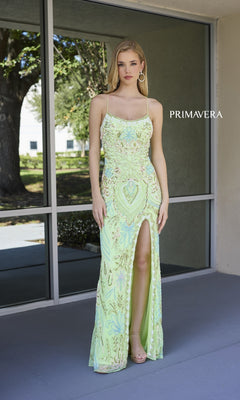A woman stands on a porch wearing the Beaded-Print Designer Prom Dress: Primavera 4156, featuring spaghetti straps and a thigh-high slit. The word "PRIMAVERA" appears on the wall behind her.