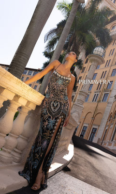 A woman in the Primavera 4156 Beaded-Print Designer Prom Dress with a high slit stands by a stone railing before a yellow building and palm trees. "PRIMAVERA" is displayed on the image.