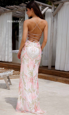 A woman stands outdoors near a white lounge area, wearing the Beaded-Print Designer Prom Dress: Primavera 4156, a floor-length backless gown with pink and gold embroidery.