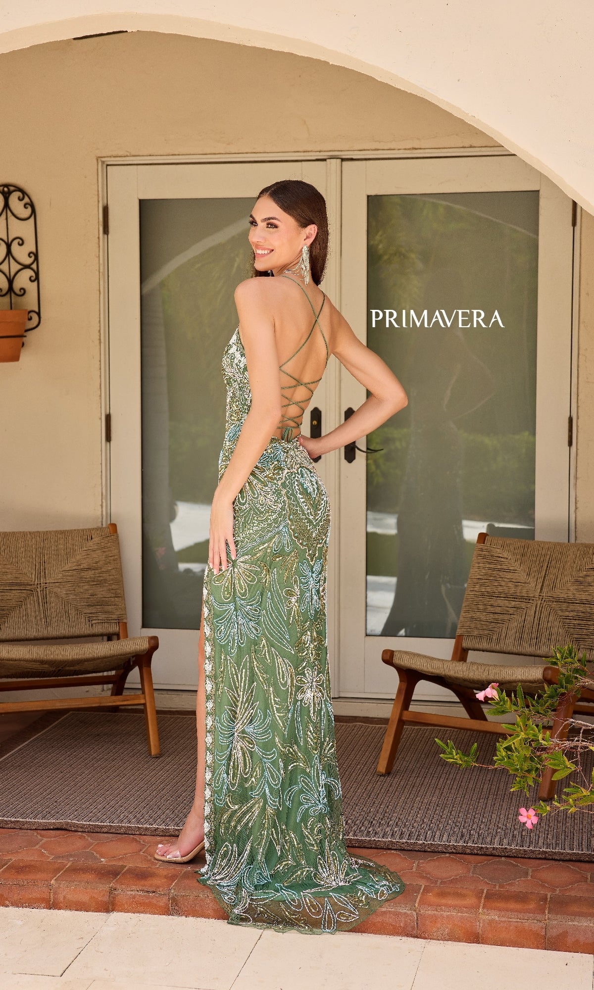 A woman stands on a patio in front of glass doors, wearing the Beaded-Print Designer Prom Dress: Primavera 4156, which features intricate beading, spaghetti straps, a backless design, and a high slit. Two wooden chairs and a 