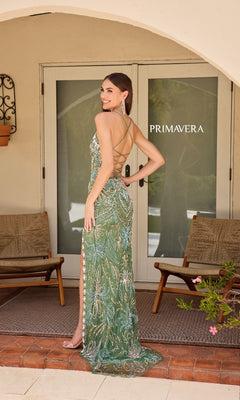 A woman stands on a patio in front of glass doors, wearing the Beaded-Print Designer Prom Dress: Primavera 4156, which features intricate beading, spaghetti straps, a backless design, and a high slit. Two wooden chairs and a "Primavera" sign are behind her.