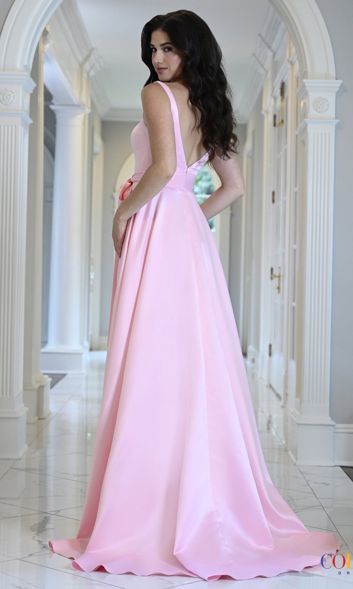 A woman wears a sleeveless pink satin A-line Long Prom Dress: Colors Dress 4157 with a v-neckline and a bow sash at the waist while standing in a hallway.