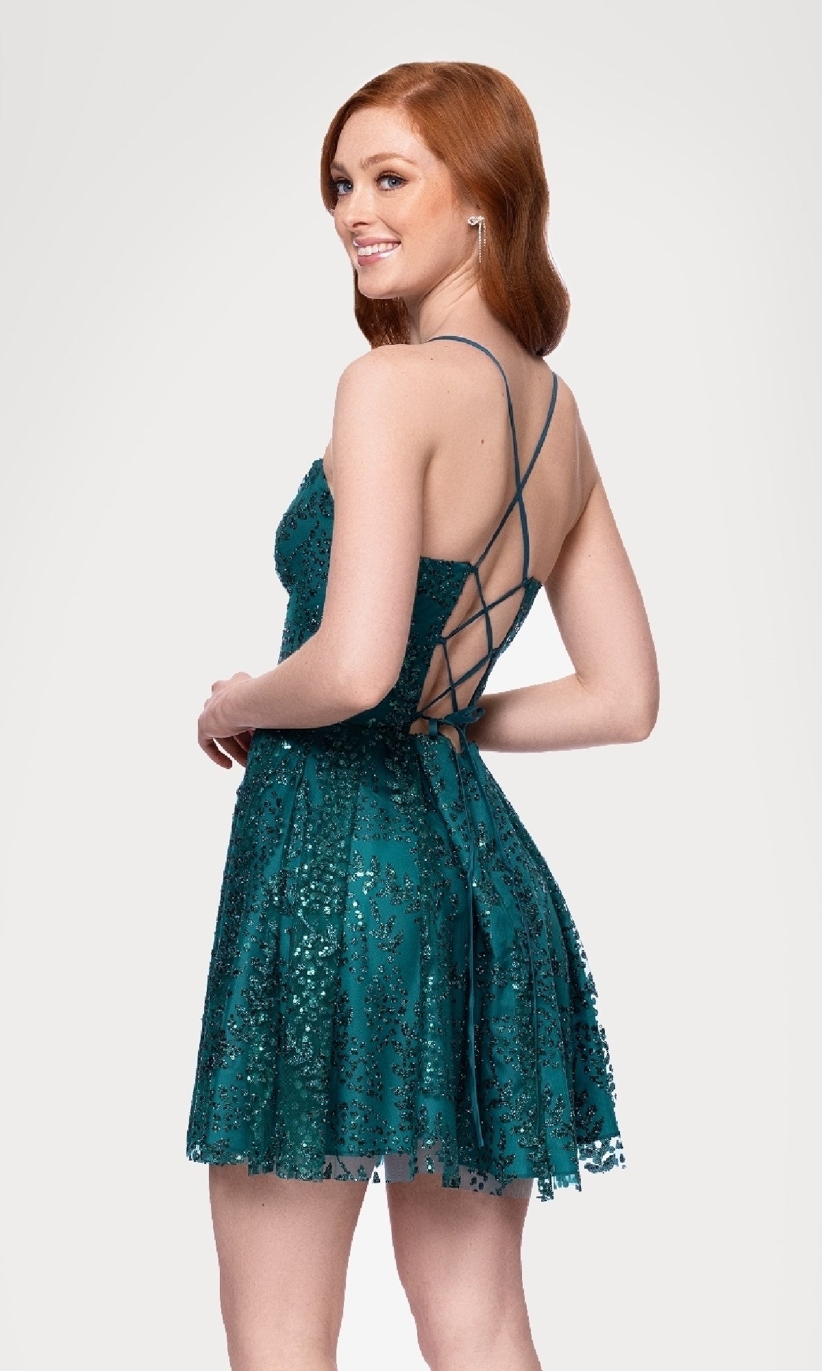 A woman with long red hair wears the Short Teal Glitter Hoco Dress (Blondie Nites 4159BN) featuring a lace-up strappy-back, smiling over her shoulder against a plain light background.