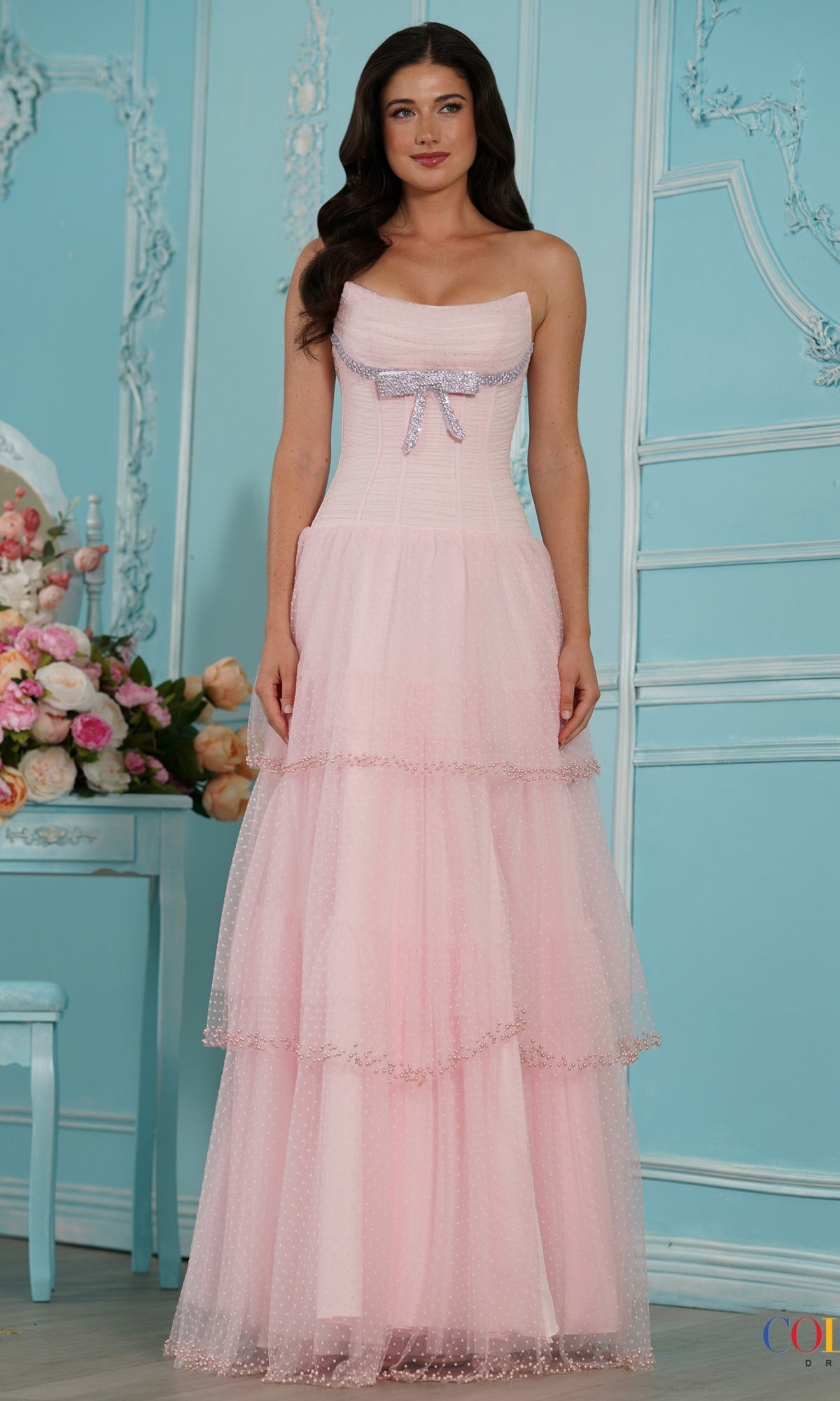 A woman wears a pink Long Prom Dress: Colors Dress 4175, featuring a strapless, tiered tulle skirt with a beaded bodice and a rhinestone bow detail.