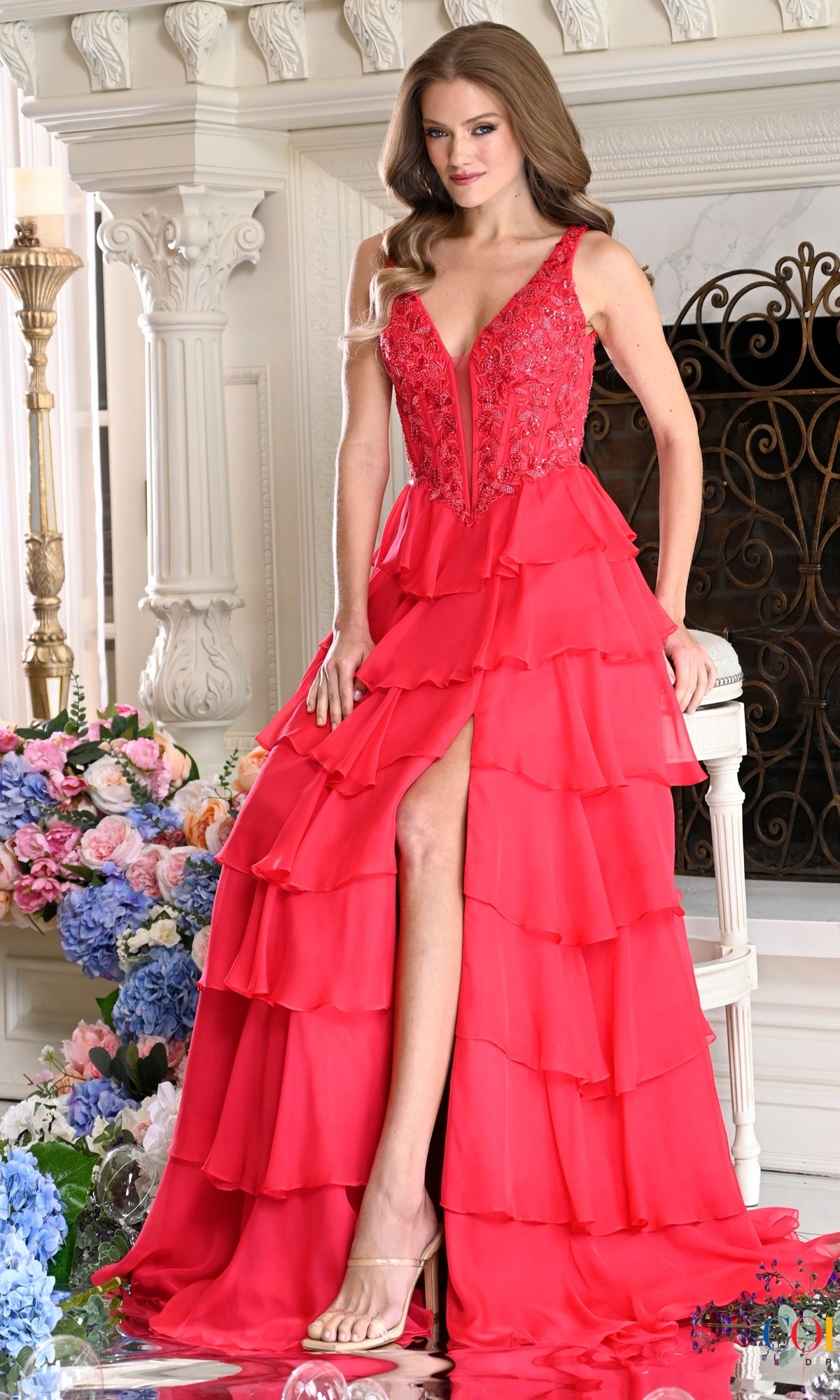 A woman wearing a watermelon-red Colors Dress 4177 V-neck prom dress with a lace bodice, a layered chiffon ruffle skirt, and a high leg slit.
