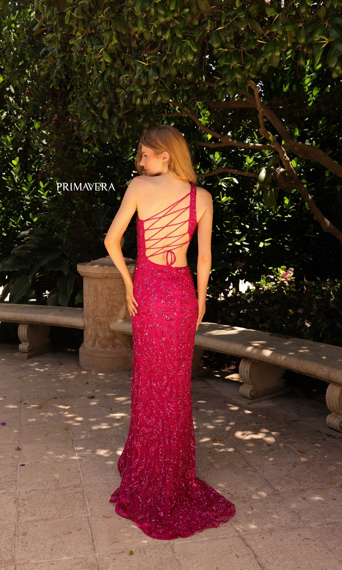 A woman stands outdoors on a stone patio near greenery, wearing the Primavera Sequin-Print Long Prom Dress 4191 in bright pink with lace-up details.