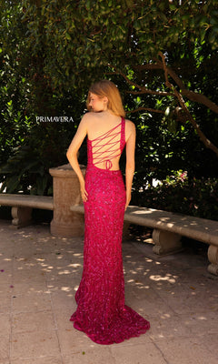 A woman stands outdoors on a stone patio near greenery, wearing the Primavera Sequin-Print Long Prom Dress 4191 in bright pink with lace-up details.