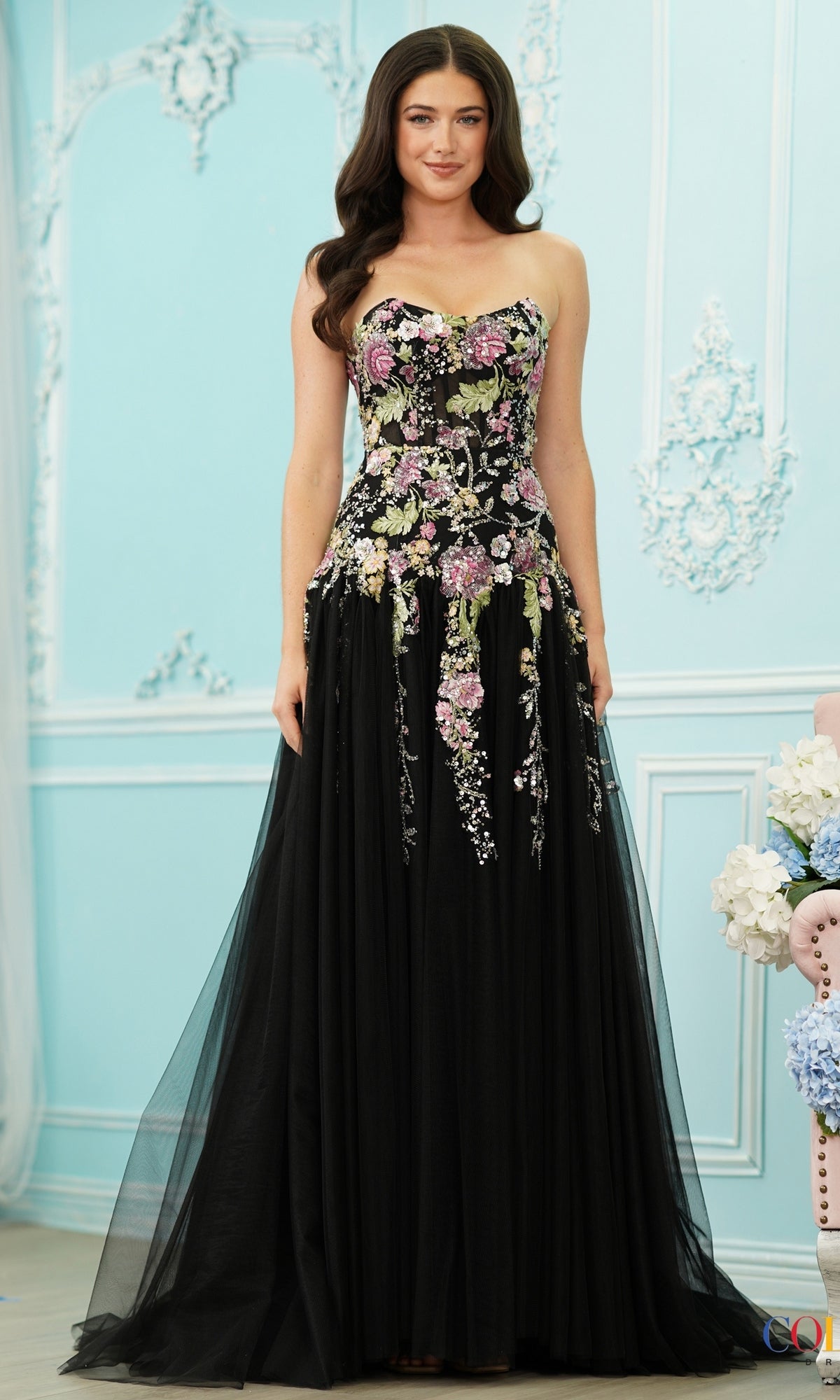 A woman wearing a Long Prom Dress: Colors Dress 4199, a strapless, floor-length black floral gown with an embroidered bodice and a full tulle skirt.