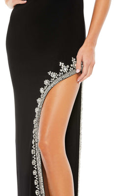 A person wearing the Mac Duggal 42002 Bead-Trimmed Long Formal Dress, featuring a high slit, black fabric, sparkling rhinestones along the edge, and a hand resting near the exposed leg.