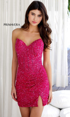 A woman wearing the Strapless Beaded Mini Hoco Dress: Primavera 4201, a bright pink sequined sweetheart mini dress with a thigh-high slit, stands indoors near white curtains. The fitted cocktail style dazzles with bold elegance.