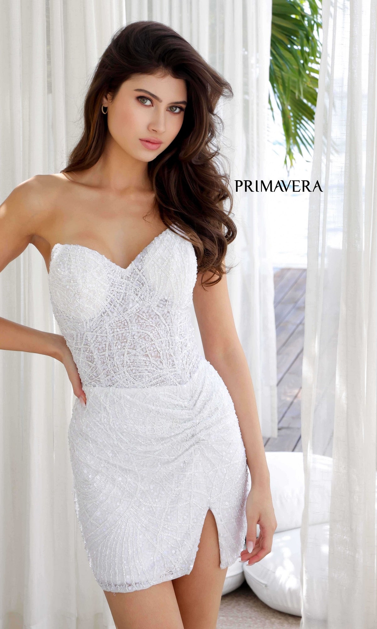 A woman poses indoors near sheer curtains in the Strapless Beaded Mini Hoco Dress: Primavera 4201, a white strapless sweetheart mini with beadwork and a front slit, with lush greenery visible through the window.