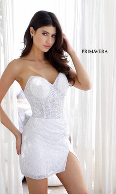 A woman in the Strapless Beaded Mini Hoco Dress: Primavera 4201 stands indoors, touching her hair. The white, textured mini shines under the light, with "PRIMAVERA" displayed on the right side of the image.
