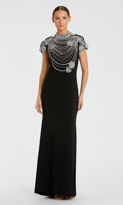 A woman wears the Mac Duggal 42055, a floor-length black formal dress with short sleeves, beaded details, and intricate silver embellishments on the shoulders and chest.